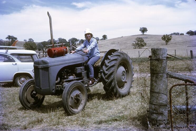 Woman smiles driving grey tractor. Gate at right, hills in background.