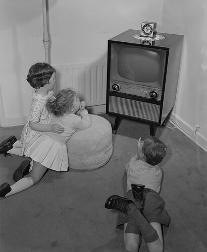 three young children watching a floor console television set in their lounge room.