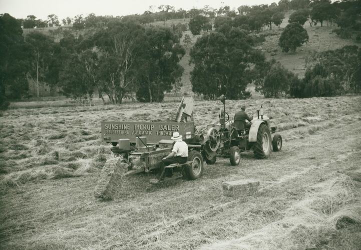 A man driving a tractor towing a Pickup Baler with a man sitting on the back operating it.