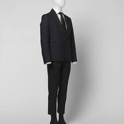Black suit made of black and grey fabric. Two pockets. White shirt, black tie. Three quarter view.