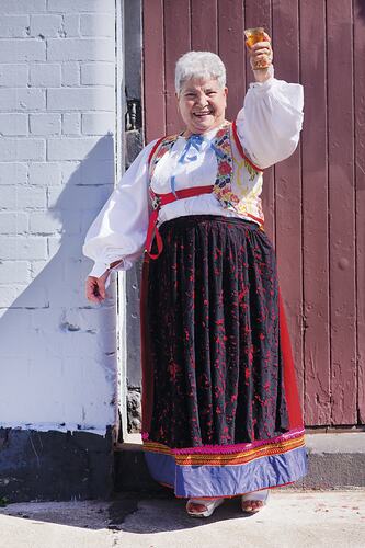 Woman in white shirt, colourful vest and black skirt with red trim holds up a cup and smiles.
