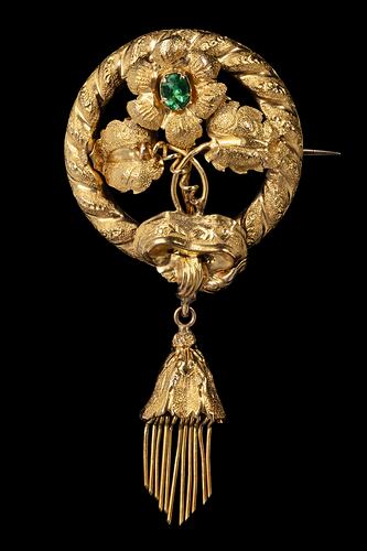 Round gold brooch. Top central flower has a green paste stone. Flower supported by two leaves and tendrils. Round border entwined with gold ribbon. Tassel hangs from base.