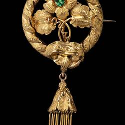 Round gold brooch. Top central flower has a green paste stone. Flower supported by two leaves and tendrils. Round border entwined with gold ribbon. Tassel hangs from base.
