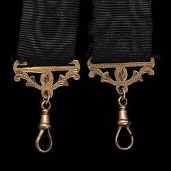 Front of detail of ends of two black ribbed straps, each with a gold decorative attachment and tear-shaped clip.