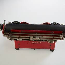 Red metal typewriter, 32 keys, black plastic spacebar at front, black roller for paper at top back. Back view.
