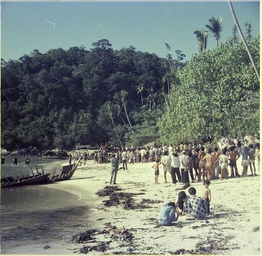 Large group of Vietnamese refugees on the beach. Trees to the right and heavily treed hill behind them.