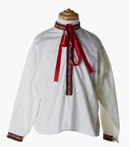 White long sleeve shirt. Red geometric patterned band embroidered at cuffs and collar. Also down front centre of shirt. Red ribbon bow at neck.