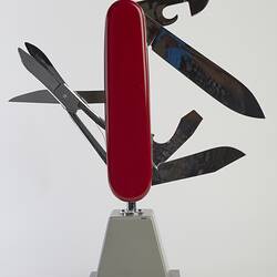 Model of muti-tool on stand. Red handle with all tools out. Large blade, short blade, scissors, bottle opener (with flathead screwdriver), a can opener (with flathead screwdriver), and a reamer.