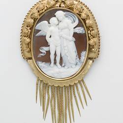 Oval cameo brooch with white winged figure and female, brown background. Ornate gold border and hanging fringe.