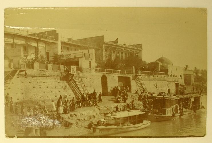 People standing along river front and on boats, buildings in background.