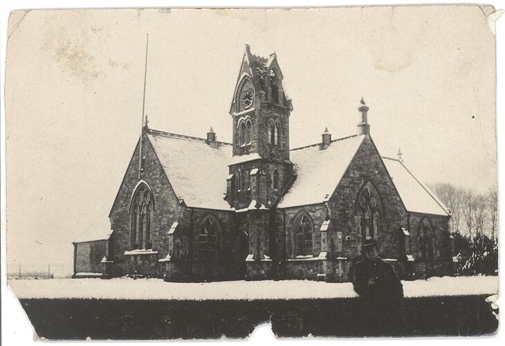 Man standing in front of hedge fence, with snow covered church building in background.