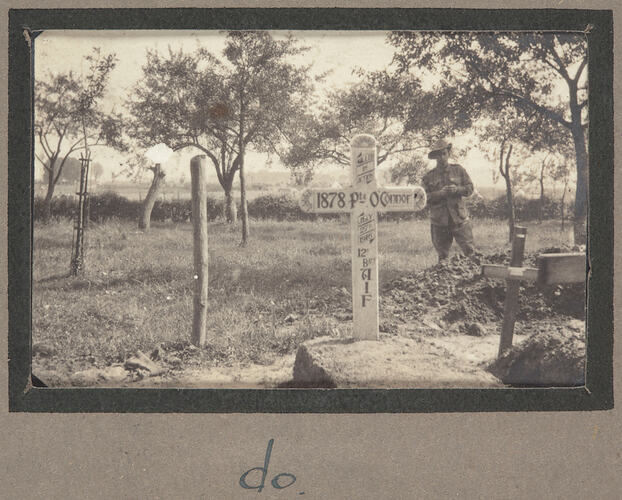 Grave with cross shaped marker covered in writing with servicemen and trees in the background.