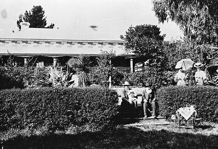 Man, woman and baby seated on steps in front of house. Hedge in foreground.