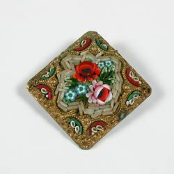 Bronze coloured diamond shaped metal brooch with plastic floral design in centre in off-white, green and red.