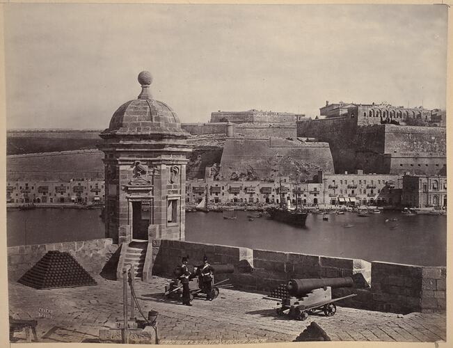 Township with fortifications in foreground and port in background. Two soldiers stand beside a canon.