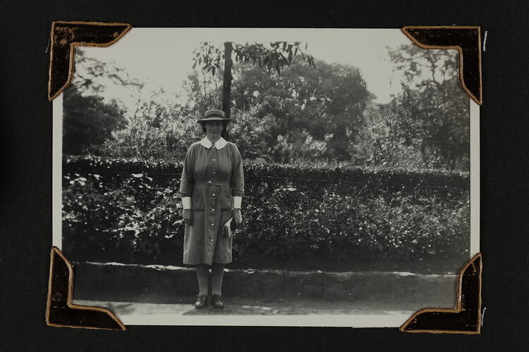 Woman in uniform standing in front tree shrubs with garden behind.