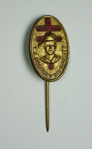 Oval-shaped, gold coloured metal lapel pin, with bust of soldier above text and red cross in background.