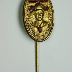 Oval-shaped, gold coloured metal lapel pin, with bust of soldier above text and red cross in background.