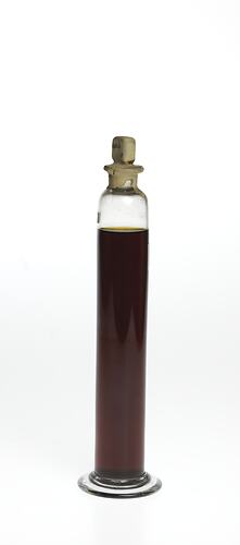 Cylindrical glass jar with dark brown liquid. One label affixed, sealed at top.