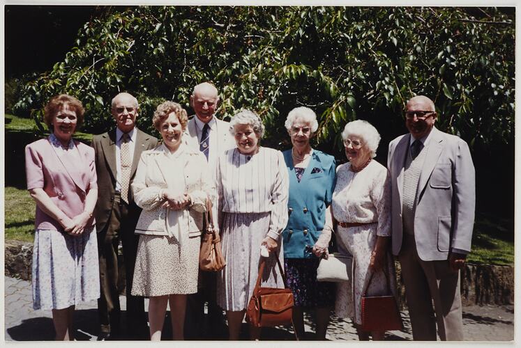 Five older women and three older men pose outdoors with trees behind them.