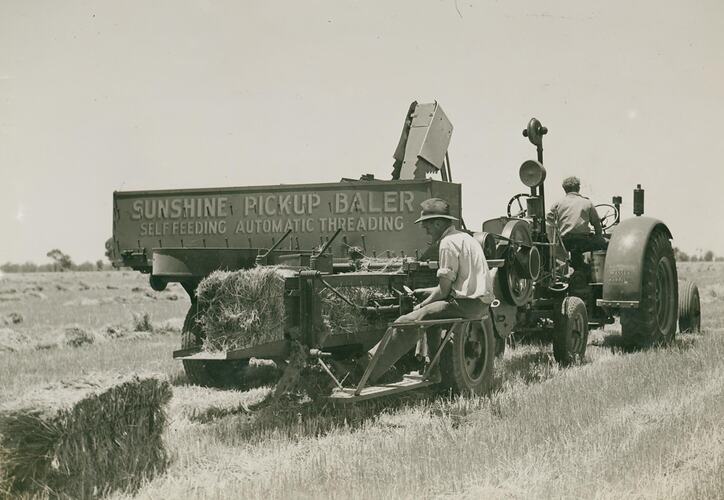Man driving a tractor towing a pickup baler, with a man sitting on the back tying knots in the wire.