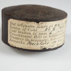 Disc-shaped dark brown briquette with flat sides. An inscribed paper band is affixed around the disc.