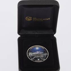 Round silver coin with runnning dinosaur. Has dark coloured background. Coin housed in dark blue velvet box.