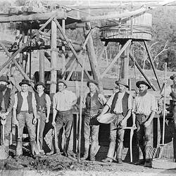 [Goldminers at Glenpatrick, near Ararat, about 1880. Behind the miners stand a poppet head and a whim for raising and lowering a pulley into the mine shaft.]