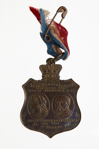 Shield-shaped medal with Queen Victoria and Edward VII faces. Crown on top. Red, yellow, blue ribbon on pin.