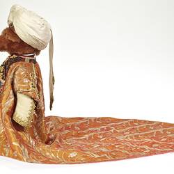 Profile of light brown plush bear wearing white turban, ornate tunic and long coat with gold embroidery.