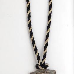 Back of shield-shaped medal with black and white twined cord. Raised text.