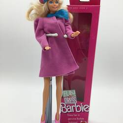 Barbie doll with blonde hair wearing pink with blue trim coat and hat. Pink Barbie box beside her.