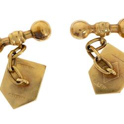 Reverse view of gold cufflinks of heraldic shield design chains to bar-bells.