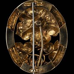 Back of gold brooch with large flower with red centre, supported by two lower leaves and tendrils, inside an oval border. Vertical pin.