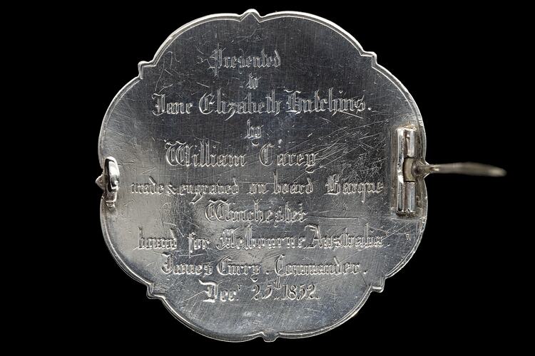 Back of brooch made from silver coin. Scrolled edge. Raised lettering. Pin across centre is open.