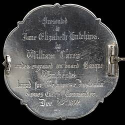 Back of brooch made from silver coin. Scrolled edge. Raised lettering. Pin across centre is open.
