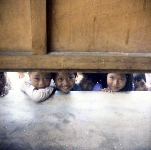 Six Vietnamese refugee children, looking under a timber wall partition.