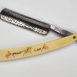 Cut throat razor, metal blade, ivory-styled plastic handle with 'PHAR LAP', picture of jockey on racehorse and two flags. Open.