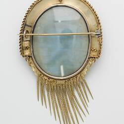 Oval cameo brooch with white winged figure and female, brown background. Ornate gold border and hanging fringe. Back view.
