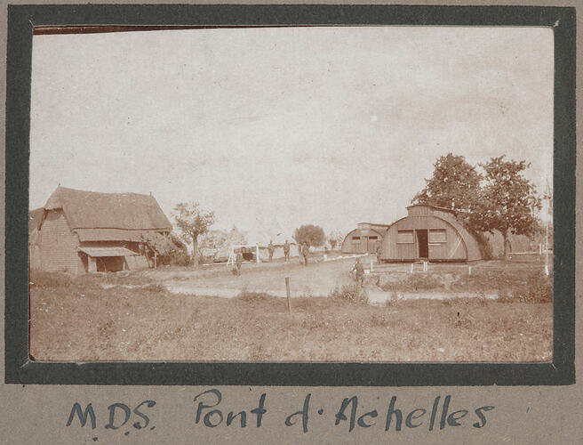 Farm house in the left, tents in the middle and two huts on right, with servicemen standing in the background.