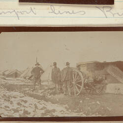 Three servicemen standing next to a horse drawn cart with tents in the background.