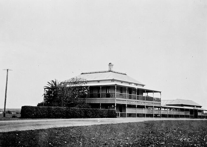 Two storey hotel with balcony beside a road. Palm tree and hedge fence in front.