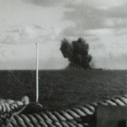 View of a bomb explosion on the ocean with billowing smoke, seen from the deck of a ship.