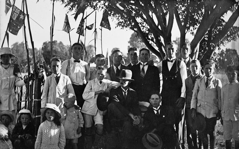 Group portrait of formally dressed men, women and children. Flags attached to long tree branches in background