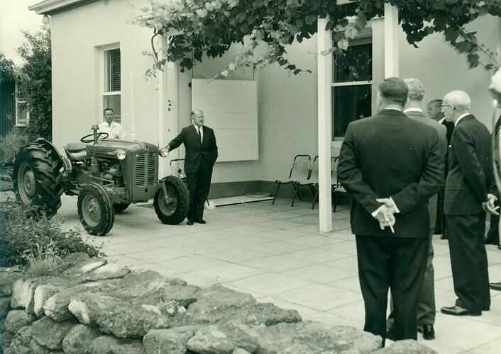 A large group of staff, stand on a verandah, looking at a  tractor.