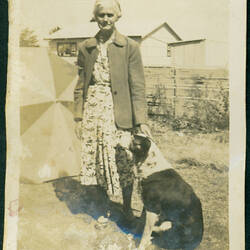 Older woman standing in garden with dog. Beach umbrella and building in background.