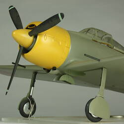 Green grey plane model with red spot on each wing and fusleage. Yellow nose, black propeller. Propeller detail.