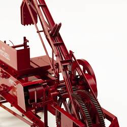 Red metal hay press model with turning gears and springs, and white painted letters on side.