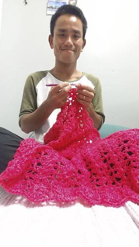 Man sitting cross-legged crocheting pink blanket.