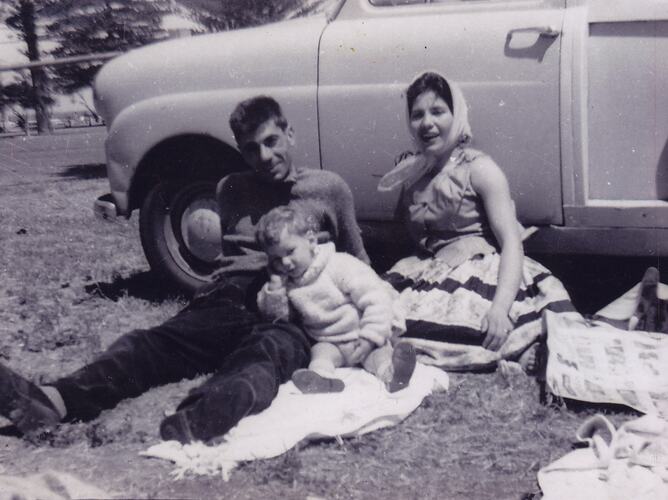 Man, woman and young boy sit on picnic rug and lean against a parked car.
